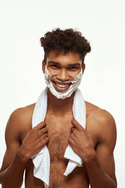 Cropped of smiling black guy with shaving foam on his face looking at camera. Young brunette curly man with towel and naked torso. Hygiene and skin care. Isolated on white background. Studio shoot - Photo, Image