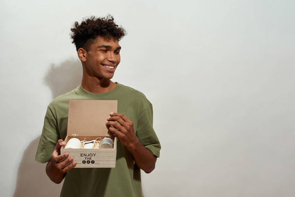 Smiling black guy holding open wooden box with set of creams and looking away. Young brunette curly man wearing t-shirt. Body and skin care. Isolated on white background. Studio shoot. Copy space - Foto, Imagen