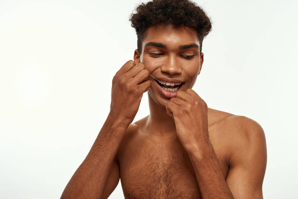 Partial of black guy brushing his teeth with dental floss. Young brunette curly man with naked torso. Dental care and hygiene. Isolated on white background. Studio shoot. Copy space - Zdjęcie, obraz