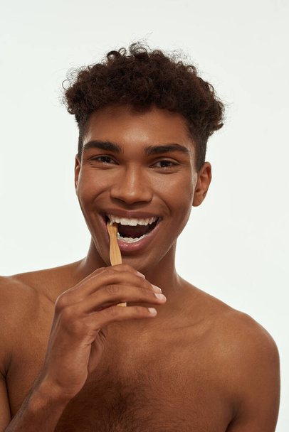 Partial of pleased black guy brushing his teeth with toothpaste. Young brunette curly man with naked torso looking at camera. Dental care and hygiene. Isolated on white background. Studio shoot - Фото, зображення