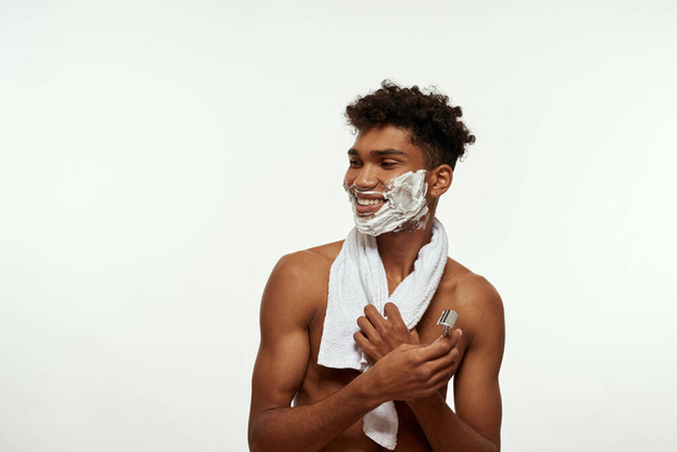 Smiling black guy with shaving foam on face holding razor and looking away. Young man with towel and naked torso. Hygiene and skin care. Isolated on white background. Studio shoot. Copy space - Foto, afbeelding