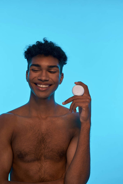 Smiling black guy holding and showing dental floss. Young brunette curly man with naked torso looking away. Dental care and hygiene. Isolated on light blue background. Studio shoot. Copy space - Photo, Image