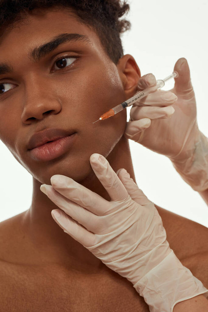 Doctor hands wearing latex gloves making injection of botox with syringe on face of black guy. Cropped young man with naked torso. Face lifting and mesotherapy. Isolated on white background in studio - Photo, image