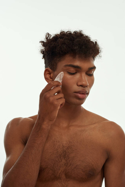 Cropped image of black guy applying ice slice on his face. Young serious slim brunette curly man with naked torso. Body and skin care. Isolated on white background. Studio shoot - Zdjęcie, obraz