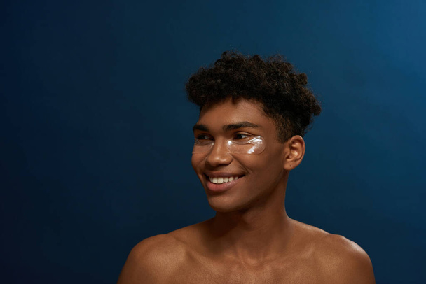 Cropped of smiling black guy with under eye patches on face look away. Young brunette curly man. Body and skin care. Facial lifting and rejuvenation. Isolated on blue background in studio. Copy space - 写真・画像