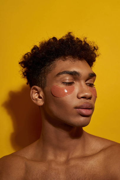 Partial image of black guy with under eye patches on face. Young brunette curly man with naked torso. Body and skin care. Facial lifting and rejuvenation. Yellow background. Studio shoot. Copy space - Foto, imagen
