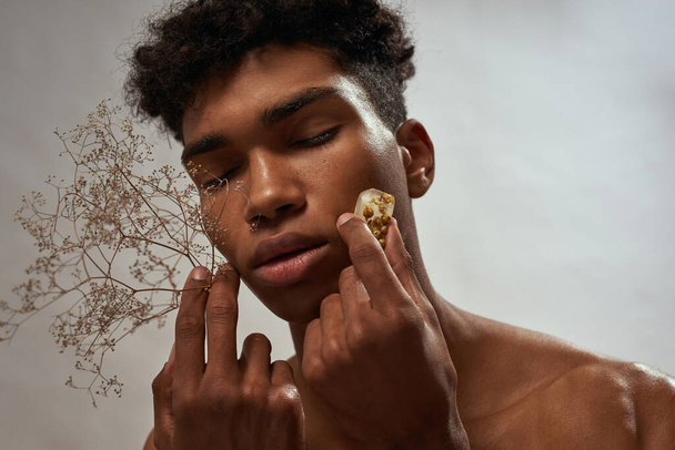 Partial of black guy applying ice slice with plant inside on his face. Young curly man with closed eyes. Body and skin care. Concept of natural cosmetic. Isolated on white brown background in studio - Photo, Image