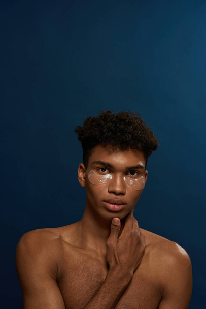 Portrait of confident black guy with under eye patches on face looking at camera. Young man with naked torso. Body and skin care. Facial lifting and rejuvenation. Blue background in studio. Copy space - Fotografie, Obrázek