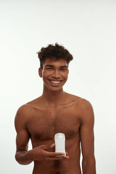 Smiling black guy holding deodorant and looking at camera. Young slim brunette curly man with naked torso. Male beauty and body care. Isolated on white background. Studio shoot. Copy space - Photo, image