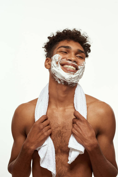 Partial of smiling black guy with shaving foam on his face. Young brunette curly man with towel and naked torso looking away. Hygiene and skin care. Isolated on white background. Studio shoot - Fotoğraf, Görsel