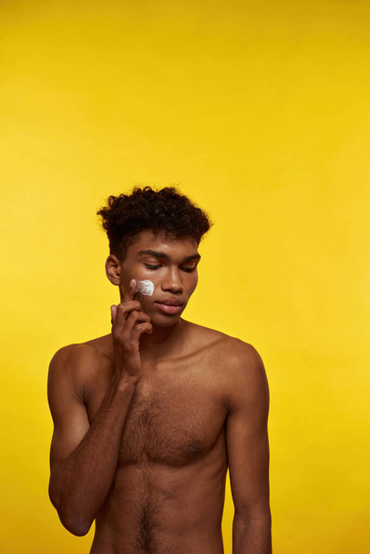 African american guy smearing cosmetic cream on his face. Young brunette curly man with naked torso. Concept of facial skin care. Isolated on yellow background. Studio shoot. Copy space - Zdjęcie, obraz