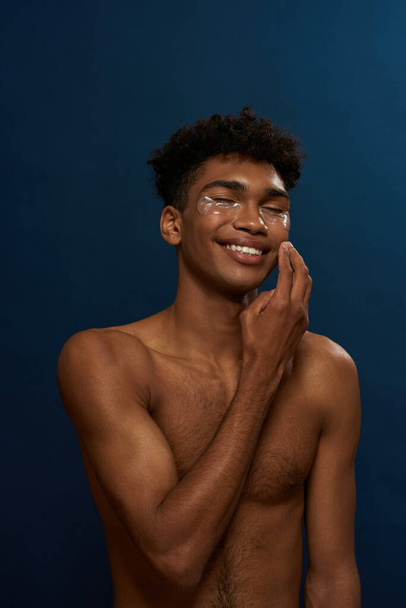 Cropped of smiling black guy correct under eye patch on face. Young man with naked torso and closed eyes. Body and skin care. Facial lifting and rejuvenation. Blue background. Studio shoot. Copy space - Foto, afbeelding