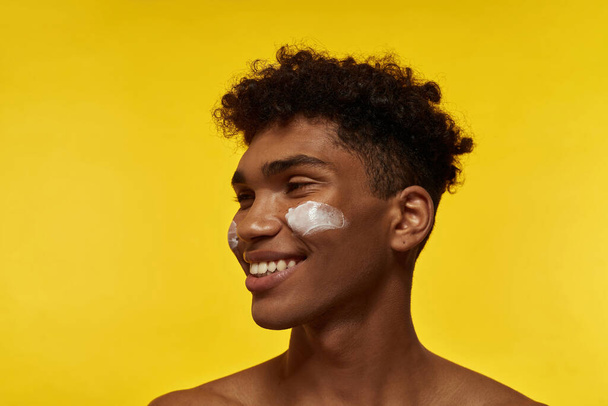 Cropped image of smiling black guy with cosmetic cream on his face looking away. Young brunette curly man. Facial skin care. Isolated on yellow background. Studio shoot. Copy space - Zdjęcie, obraz