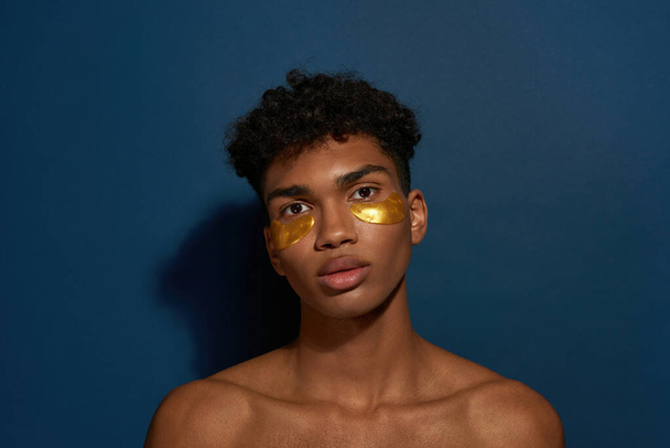 Portrait of serious black guy with under eye patches on face. Young man with naked torso looking at camera. Body and skin care. Facial lifting and rejuvenation. Blue background. Studio shoot. - Фото, изображение