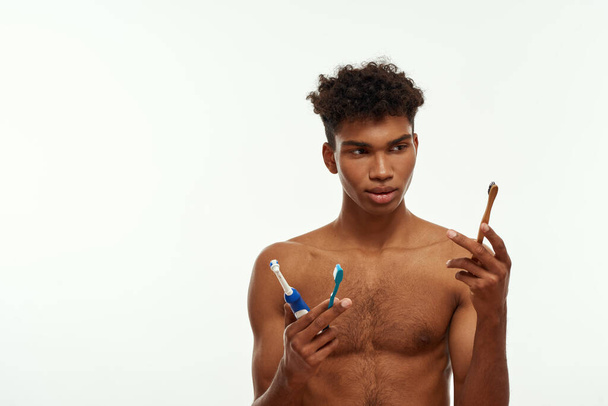 Concentrated black guy looking at toothbrush from three kinds in his hands. Young brunette curly man with naked torso. Dental care and hygiene. Isolated on white background. Studio shoot. Copy space - Foto, Bild