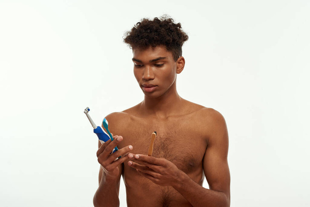 Focused black guy choosing and looking at toothbrushes from three kinds. Young brunette curly man with naked torso. Dental care and hygiene. Isolated on white background. Studio shoot. Copy space - Фото, изображение