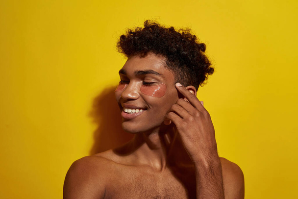 Partial image of smiling black guy with under eye patch on face. Young man with naked torso. Body and skin care. Facial lifting and rejuvenation. Isolated on yellow background in studio. Copy space - Valokuva, kuva