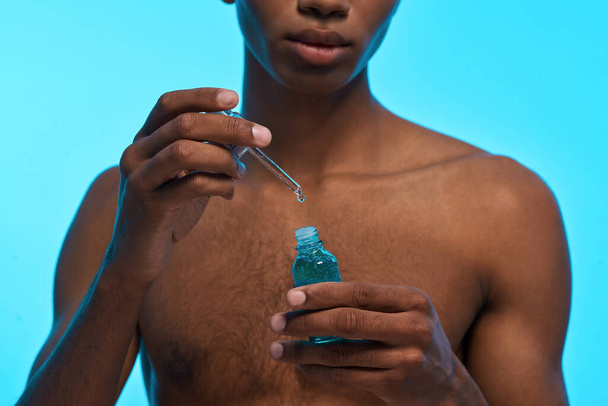 Partial image of black guy holding dropper and bottle with natural organic cosmetic oil. Obscure face of young man with naked torso. Body and skin care. Isolated on light blue background. Studio shoot - Фото, зображення