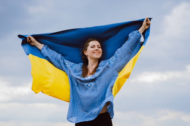 Happy free ukrainian woman with national flag on dramatic sky background. Portrait of lady in blue embroidery vyshyvanka shirt. Ukraine, independence, patriot symbol. High quality photo - Photo, Image