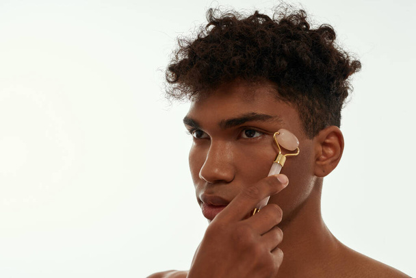 Partial image of black guy massaging his face with jade roller. Young concentrated brunette curly man with naked torso. Body and skin care. Isolated on white background. Studio shoot. Copy space - Φωτογραφία, εικόνα