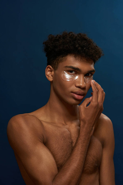 Partial of black guy apply under eye patch on face. Young serious man with naked torso looking at camera. Body and skin care. Facial lifting and rejuvenation. Blue background. Studio shoot. Copy space - Photo, Image