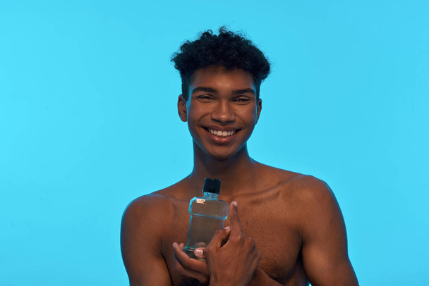 Smiling african american guy holding and pointing with finger at bottle with shower gel. Young man with naked torso. Hygiene and body care. Isolated on light blue background. Studio shoot. Copy space - Фото, зображення