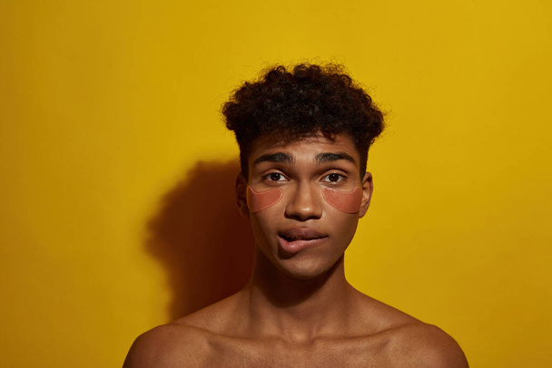 Partial of worried black guy with under eye patches on face biting his lip. Young man look at camera. Body and skin care. Facial lifting and rejuvenation. Yellow background. Studio shoot. Copy space - Photo, image