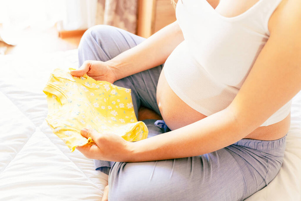 Детская одежда беременной женщины. Красивая беременная мать с желтой детской одеждой. Беременная женщина обнимает живот и собирает сумку из роддома. Концепция материнства, беременности, родов - Фото, изображение