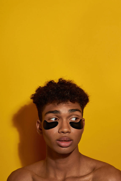 Cropped of focused black guy with under eye patches on face looking away. Young brunette curly man. Body and skin care. Facial lifting and rejuvenation. Yellow background. Studio shoot. Copy space - Φωτογραφία, εικόνα