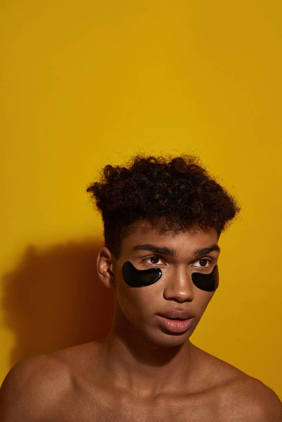 Partial of focused black guy with under eye patches on face looking away. Young man with naked torso. Body and skin care. Facial lifting and rejuvenation. Yellow background. Studio shoot. Copy space - Фото, изображение
