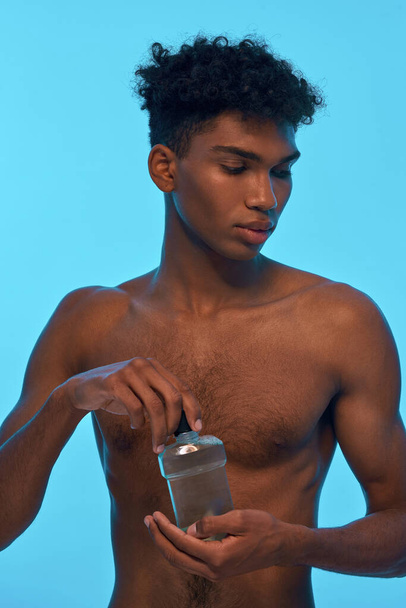 Thoughtful black guy opening bottle with shower gel. Young slim brunette curly man with naked torso. Concept of hygiene and body care. Isolated on light blue background. Studio shoot - Foto, immagini
