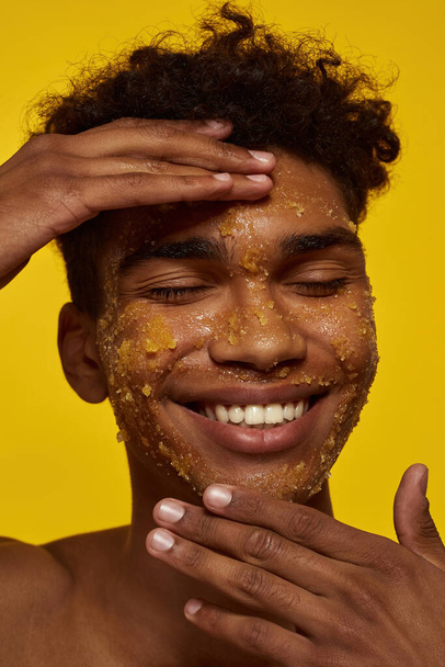 Cropped of smiling african american guy touching his face with honey scrub. Young brunette curly man with closed eyes. Facial skin care. Isolated on yellow background. Studio shoot - Foto, imagen