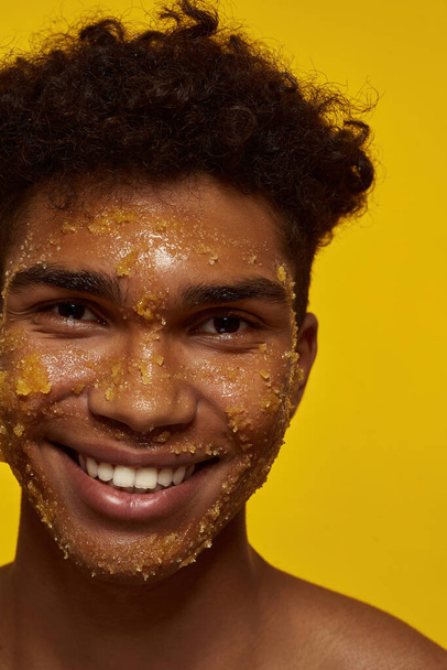 Cropped of smiling african american guy with honey scrub on his face skin looking at camera. Young brunette curly man. Facial skin care. Isolated on yellow background. Studio shoot - Photo, Image