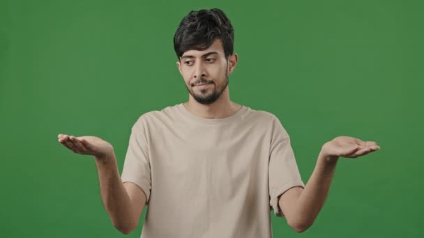 Puzzled man confused uncertain hispanic arab thoughtful young guy standing in green studio shrugs shoulders make choice decision choosing doing weights gesture with hands compares between two options - Felvétel, videó