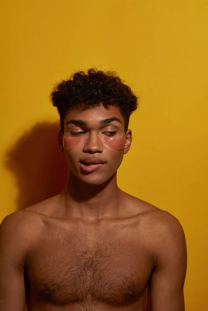 Front view of cunning black guy with under eye patches on face biting lip. Young man with naked torso. Body and skin care. Facial lifting and rejuvenation. Yellow background. Studio shoot. Copy space - Zdjęcie, obraz