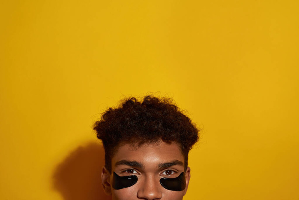 Obscure face of black guy with under eye patches on face. Young brunette curly man looking at camera. Body and skin care. Facial lifting and rejuvenation. Yellow background. Studio shoot. Copy space - Фото, зображення