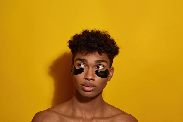 Cropped of concentrated black guy with under eye patches on face looking away. Young brunette man. Body and skin care. Facial lifting and rejuvenation. Yellow background. Studio shoot. Copy space - Foto, Imagen