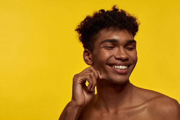 Partial image of smiling black good-looking guy with closed eyes. Young brunette curly model man. Concept of modern male beauty. Isolated on yellow background. Studio shoot. Copy space - 写真・画像