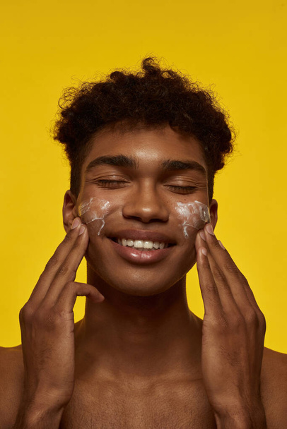 Cropped of smiling african american guy smearing cosmetic cream on his face. Young brunette curly man with closed eyes. Facial skin care. Isolated on yellow background. Studio shoot - Φωτογραφία, εικόνα
