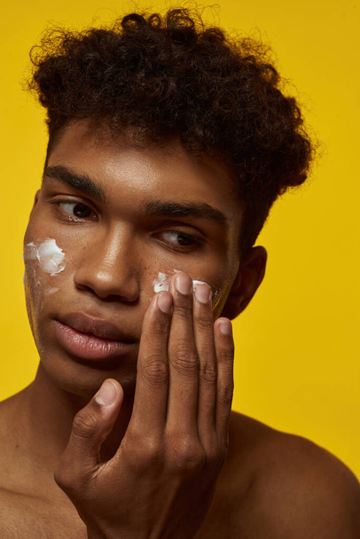 Cropped image of serious black guy smearing cosmetic cream on his face. Young brunette curly man with clean face. Concept of facial skin care. Isolated on yellow background. Studio shoot - Fotó, kép