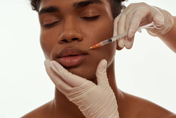 Doctor hands wearing latex gloves making injection of botox with syringe on face of black guy. Partial young man with closed eyes. Face lifting and mesotherapy. Isolated on white background in studio - 写真・画像