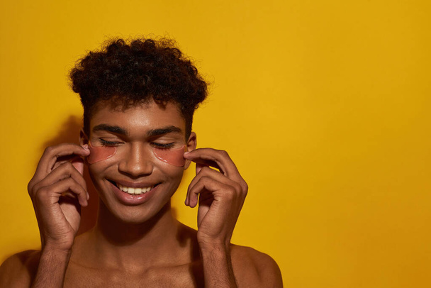 Cropped of smiling black guy applying or correcting under eye patches on face. Young brunette curly man. Body and skin care. Facial lifting and rejuvenation. Yellow background in studio. Copy space - Foto, Imagem