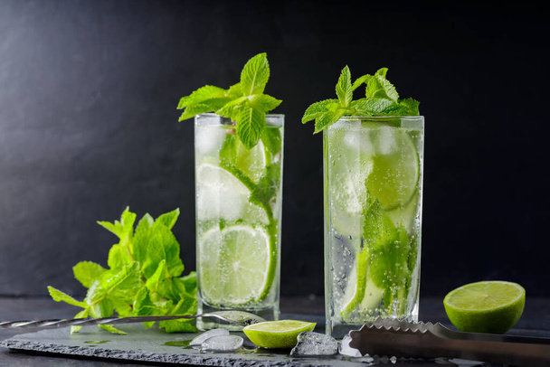 Mojito cocktail, mint, lime slices and ice cubes on a dark background. Cocktail mojito, ice tongs and cocktail spoon on slate board. Copy space - Photo, Image