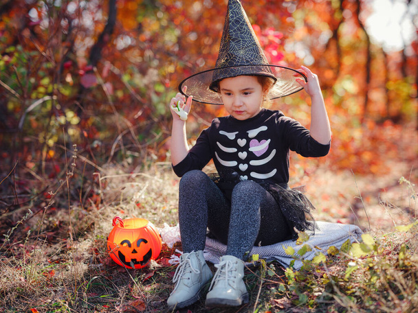 Siyah kostümlü genç kız şeker toplamaya gidiyor. Küçük cadı, balkabağı fenerli çocuklar. Sonbahar ormanında elinde şeker kovası olan çocuklar. Mutlu Cadılar Bayramı. - Fotoğraf, Görsel