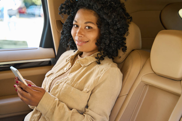 Schattig gemengd ras vrouw reist comfortabel in de achterbank van een auto, ze heeft een telefoon in haar handen - Foto, afbeelding