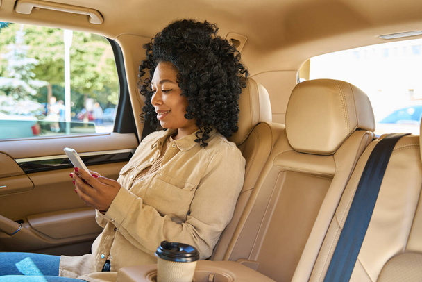 Jonge mooie passagier zit comfortabel op de achterbank van een comfortabele auto met een glas koffie op haar telefoon - Foto, afbeelding