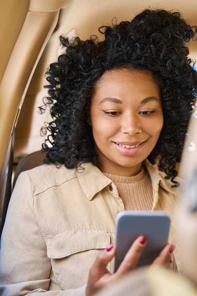 Curly-haired young mixed race female sits comfortably in passenger seat of car and looks into the phone with a smile - 写真・画像