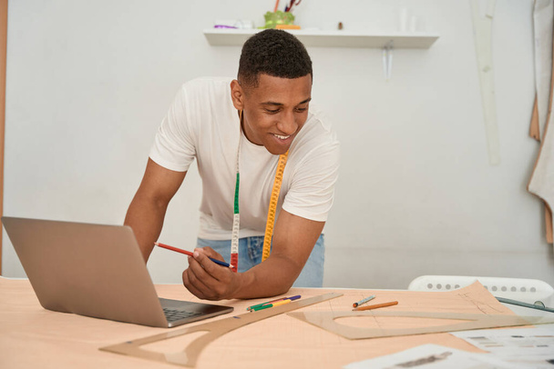 Smiling young man is standing at table with laptop and working with pencils and paper for making dress drawings - Photo, Image