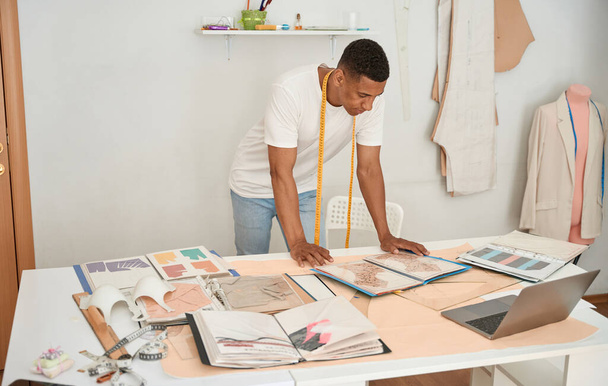Male fashion designer is standing at desk with notebook and looking at sketchbooks with dress patterns - Fotoğraf, Görsel