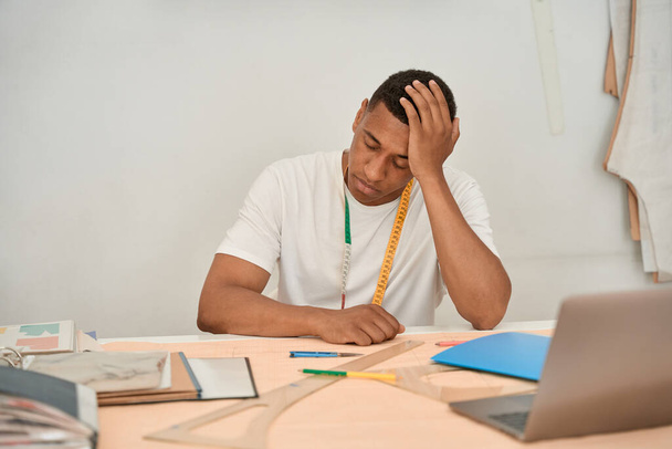 Erkek terzi, dizüstü bilgisayardan önce elinde çizimler ve kalemlerle masada oturuyor. - Fotoğraf, Görsel